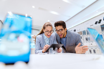 Obraz premium Cute multicultural couple in formal wear smiling and looking for new tablet to buy. Tech store interior.