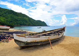 A rustic native Tobago wooden fishing boat propped up on a sandy beach by a drying net with orange floats and yellow rope, with others at anchor before lush rugged mountains under a cloudy blue sky.