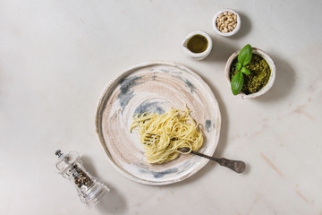 Classic italian spaghetti pasta with pesto sauce, pine nuts, olive oil and fresh basil. Served in ceramic plate with fork and ingredients above over white marble background. Flat lay, space