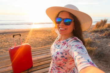Holiday, travel and tourism concept - Beautiful young woman with red suitcase taking selfies on the beach
