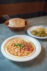 Creamy hummus with whole cheakpeas close up, pile of pitas and pickles on the background