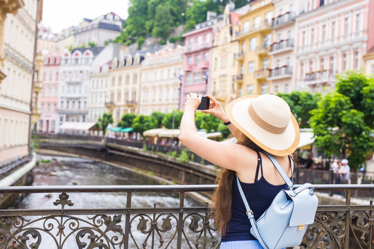 Woman taking pictures with smartphone. Stylish summer traveler woman in hat with camera outdoors in european city, old town Karlovy Vary in the background, Czech Republic, Europe.