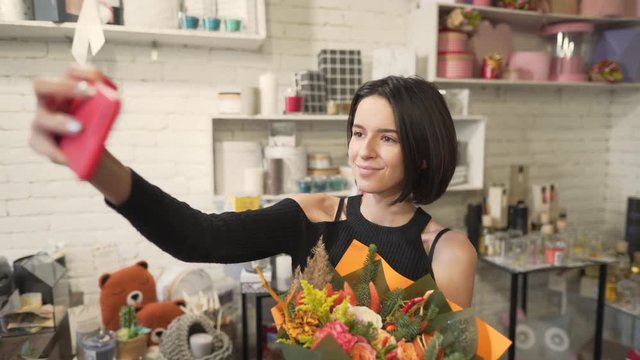 Young girl is in a flower shop. She  hold a boquet of flowers in hands and mke selfie with it.