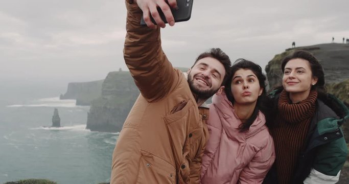 Good looking friends multi ethnic taking pictures on the top of Cliffs of Moher using a smartphone , they make funny faces and smiling large , taking video closeup.