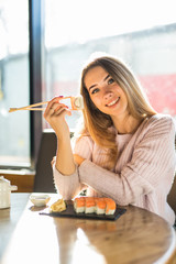 Young happy smiling pretty woman eating sushi with stick in restaurant