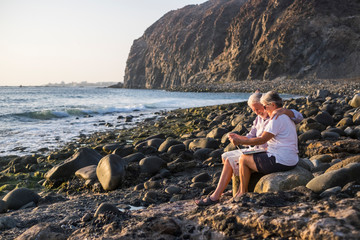 concept of vacation, technology, tourism, travel and people - happy senior couple with tablet pc computer on pebble beach with blue sea background. White hair