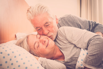 Couple of senior caucasian man and woman kissing in early morning at the bed at home for love everyday with no limit age - foreverness together concept for happy cheerful people in relationship