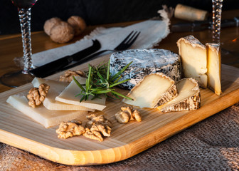 Some kinds of the blue cheeses with halves of the walnuts and rosemary, two glasses with red wines, knife and fork on the wooden desk