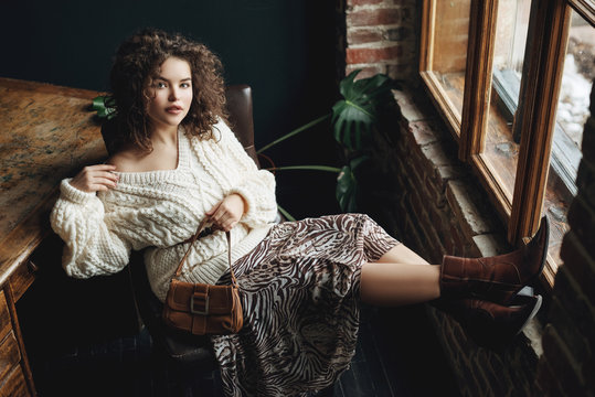 Indoor full-length fashion portrait of young beautiful confident woman wearing cozy white knitted sweater, animal print skirt, cowboy style boots, holding small brown bag, posing at loft interior
