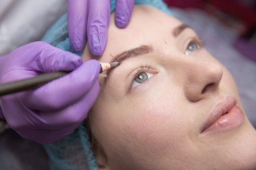 Permanent make-up for eyebrows of beautiful woman with thick brows in beauty salon. Closeup beautician doing tattooing eyebrow.