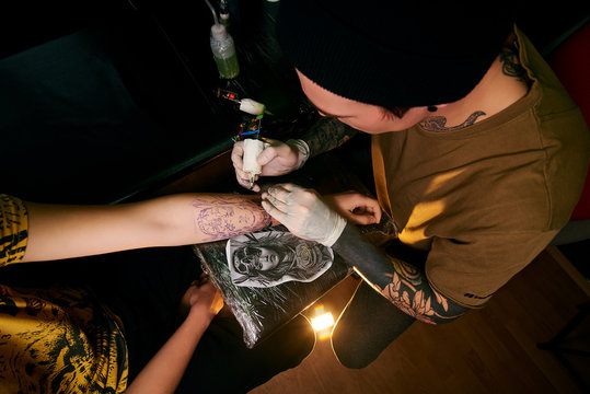 Handsome young guy in a black hat and with tattoos, beats a tattoo on his arm, tattoo salon, tattoo artist