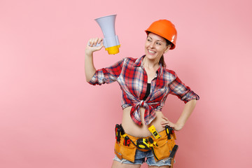 Excited handyman woman in plaid shirt, denim shorts, helmet, kit tools belt full of variety instruments hold megaphone, screaming isolated on pink background. Female in male work. Renovation concept.