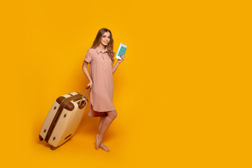 Portrait of a cheerful young woman dressed in summer clothes holding passport with flying tickets while standing with a suitcase and pointing finger away isolated over yellow background.