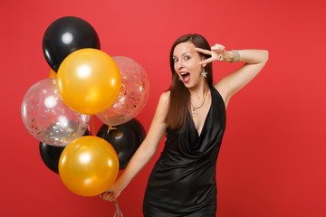 Fototapeta na wymiar Excited young girl in little black dress celebrating, showing victory sign, holding air balloons isolated on red background. St. Valentine's Day, Happy New Year, birthday mockup holiday party concept.
