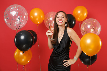 Joyful young girl in little black dress celebrating holding glass of champagne on bright red background air balloons. St. Valentine's, Women's Day Happy New Year birthday mockup holiday party concept.