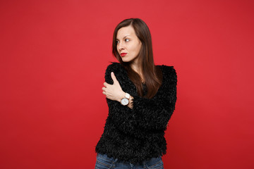 Fototapeta premium Portrait of pensive young woman in black fur sweater holding hands crossed, looking aside isolated on bright red background in studio. People sincere emotions, lifestyle concept. Mock up copy space.