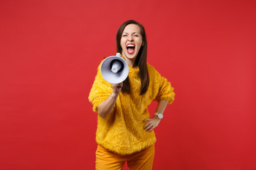 Portrait of crazy young woman in yellow fur sweater standing, screaming on megaphone isolated on...