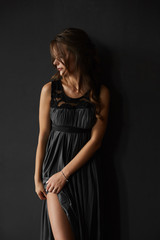 Fototapeta premium Beautiful and sensual brunette model girl with perfect body in fashionable long dress bares her long sexy legs and posing at the dark background at interior