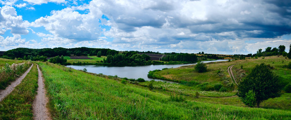 Cloudy windy summer panoramic landscape with country road and small river.Dark stormy clouds in dramatic overcast sky.Fields,green meadows and woods before the rain and storm.Rainy season.