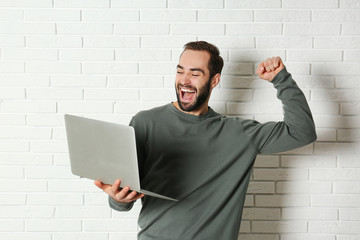 Fototapeta premium Emotional young man with laptop celebrating victory near brick wall