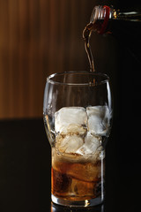 Pouring cola from bottle into glass with ice cubes on table against blurred background