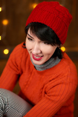 Beautiful woman is posing on dark wooden background, christmas lights and flags, holiday concept. Dressed in red knitted hat and sweater.