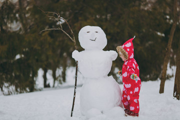 Funny little girl in winter warm clothes with hood playing, standing near snowman in park or forest...