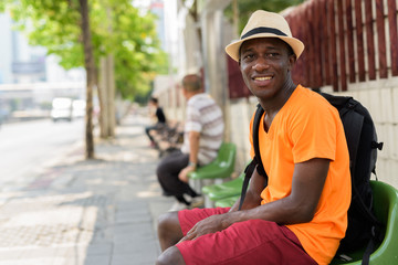 Young happy black African tourist man sitting at bus stop