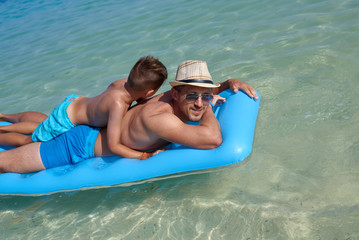 Father and son are swimming on the blue air mattress in the crystal ocean water. They are enjoying their summer holidays.