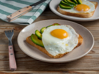 Toasts bread with fried eggs, avocado and cucumber slices on wooden table with fork and knife. Soft focus.