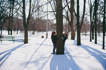 Cheerful young couple in love woman and man in warm clothes having fun near tree, walking in snowy park or forest outdoors. Winter fun, leisure on holidays. Love relationship people lifestyle concept.