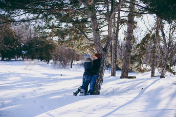 Young couple woman, man in warm clothes hugging, looking at each other near tree, walking in snowy park or forest outdoors. Winter fun, leisure on holidays. Love relationship people lifestyle concept.