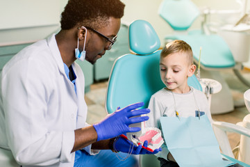 African American Man dentist in a uniform. Plastic jaw in the dentist's office. Dendist holds in his hands dental implants and talking to a little boy.