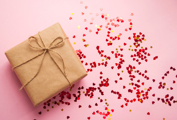 Gift box wrapped in brown colored craft paper and tied with rope on pink background with heart shape red confetti.
