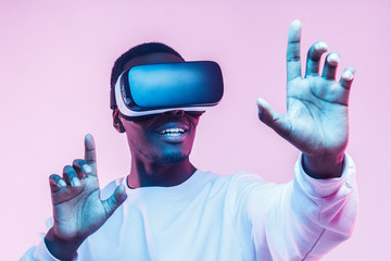 Young african man wearing  virtual reality goggles with hands up, isolated on pink background