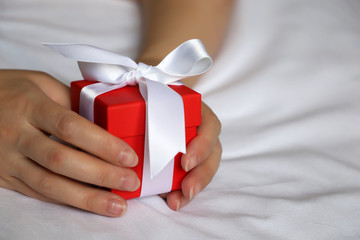 Red gift box in female hands close up. Woman on the bed holding Valentine gift tied with white ribbon, concept of birthday, romantic morning, surprise