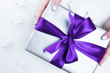 Woman's hands holding gift with atlas violet ribbon and bow on a light background with holidays decor. Holiday concept.