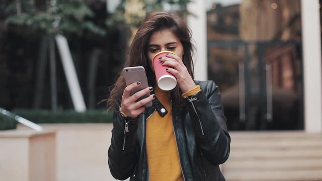 Young happy woman using smartphone and drinking coffee to go outdoors, walking down the city street, slow motion