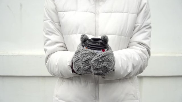 Woman warming up hands in grey woolen gloves and cup of coffee or tea in winter on white background take away
