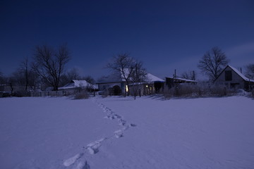 Night landscape of a snow-covered field. In the distance is seen the village. There are the footprints on the snow.