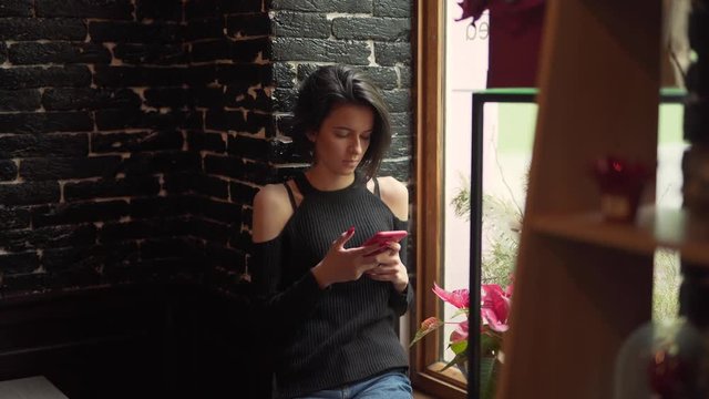 Girl is sitting near window. She hold phone in hand, chek news and send messages.