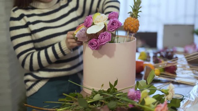 Big and beautiful box with is standing on the table. Girl is making a composition with  flowers and fruits she put them in this box making a bouget.