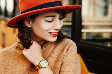 Close up outdoor portrait of young fashionable happy smiling woman wearing golden wrist watch, orange hat, beige turtleneck sweater. Copy, empty space for text