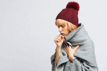 Cute sick young teen woman in red hat, wrapped in warm scarf coughing, closed eyes.Female feeling the first symptoms of illness.Bronchitis, upper respiratory infection concept.Moist cough, laryngitis