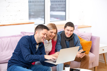 Friends sit on the couch, chatting and using a smartphone, desktop computer, laptop.