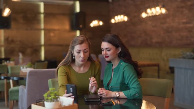Two beautiful young women are sitting in a cafe and talking in a video chat by phone. Both women are absolutely calm and relaxed. The have a conversation with a client or business partner.