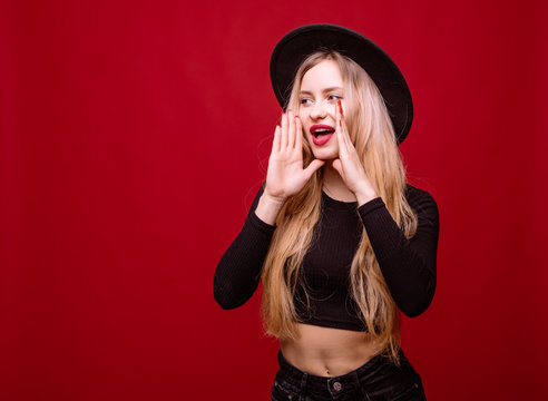 Young blonde woman in fluppy hat and black clothes scream isolated on red background