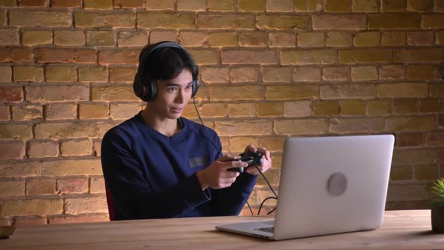 Closeup portrait of young attractive korean man in headphones playing video games on the console using the laptop
