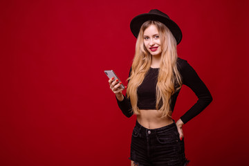 Young blonde woman in fluppy black hat and black clothes use the phone isolated on red background