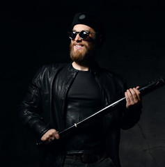 Brutal man in leather jacket, black cap and round sunglasses holds baseball bat and smiles predatory 